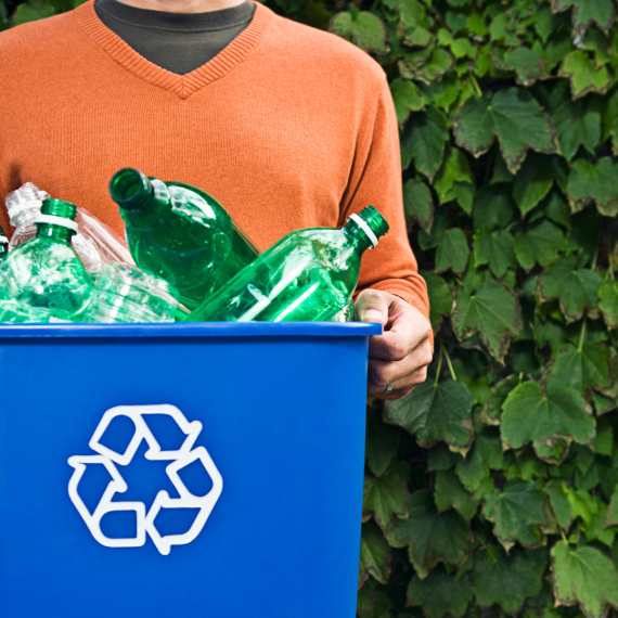 A person in an orange sweater holds a recycling bin filled with green plastic bottles.